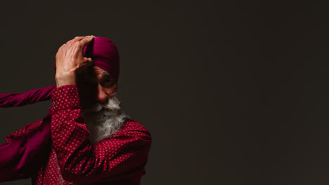 Fotografía-De-Estudio-Con-Iluminación-Tenue-De-Un-Hombre-Sikh-Mayor-Con-Barba-Atando-Una-Tela-Como-Turbante-Contra-Un-Fondo-Oscuro-Mirándose-En-El-Espejo